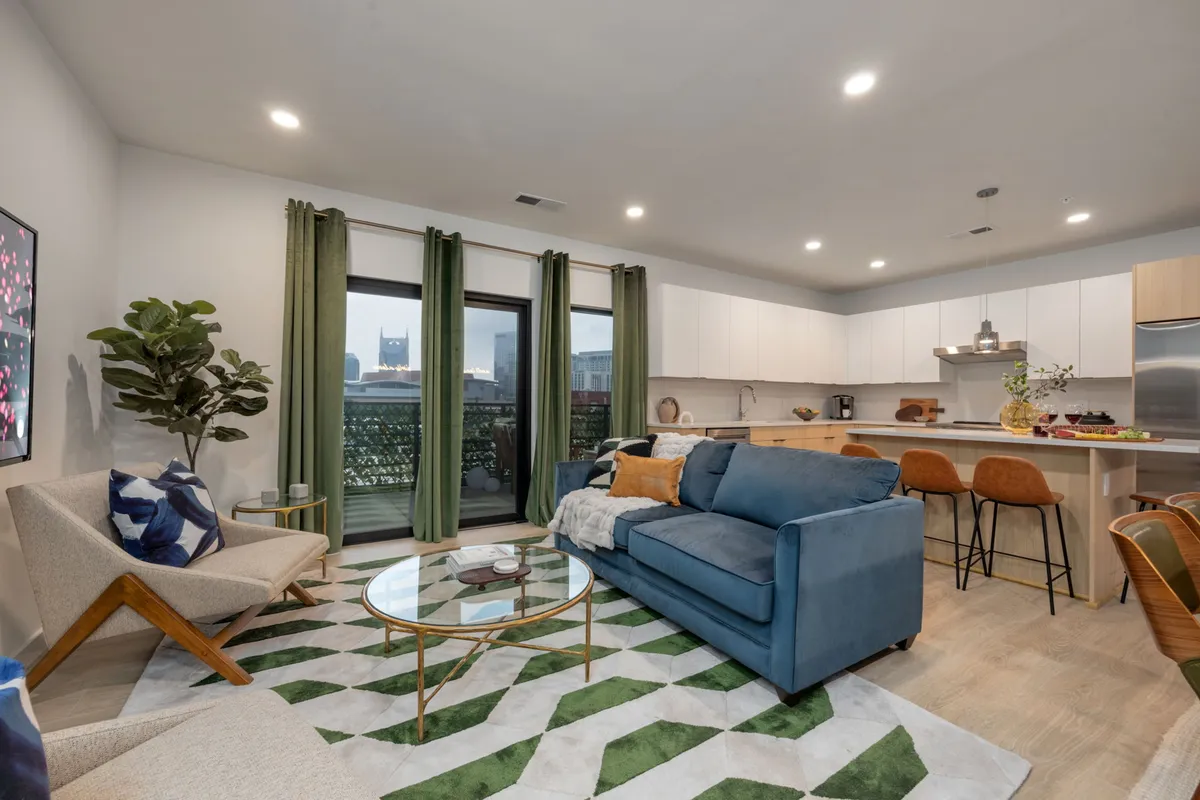 The image showcases a stylish, contemporary living area with a plush blue sofa and glass coffee table set against vibrant green curtains. Large windows frame a stunning city view of Nashville, while the open-concept kitchen area invites guests to gather and entertain. The space is sunlit and welcoming, embodying both comfort and chic design for group getaways.