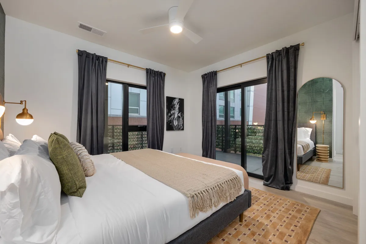 A serene bedroom featuring a bed with decorative pillows, a large window allowing natural light to pour in, and direct access to a balcony. A mirror reflects the light, making the room feel even more spacious and inviting.
