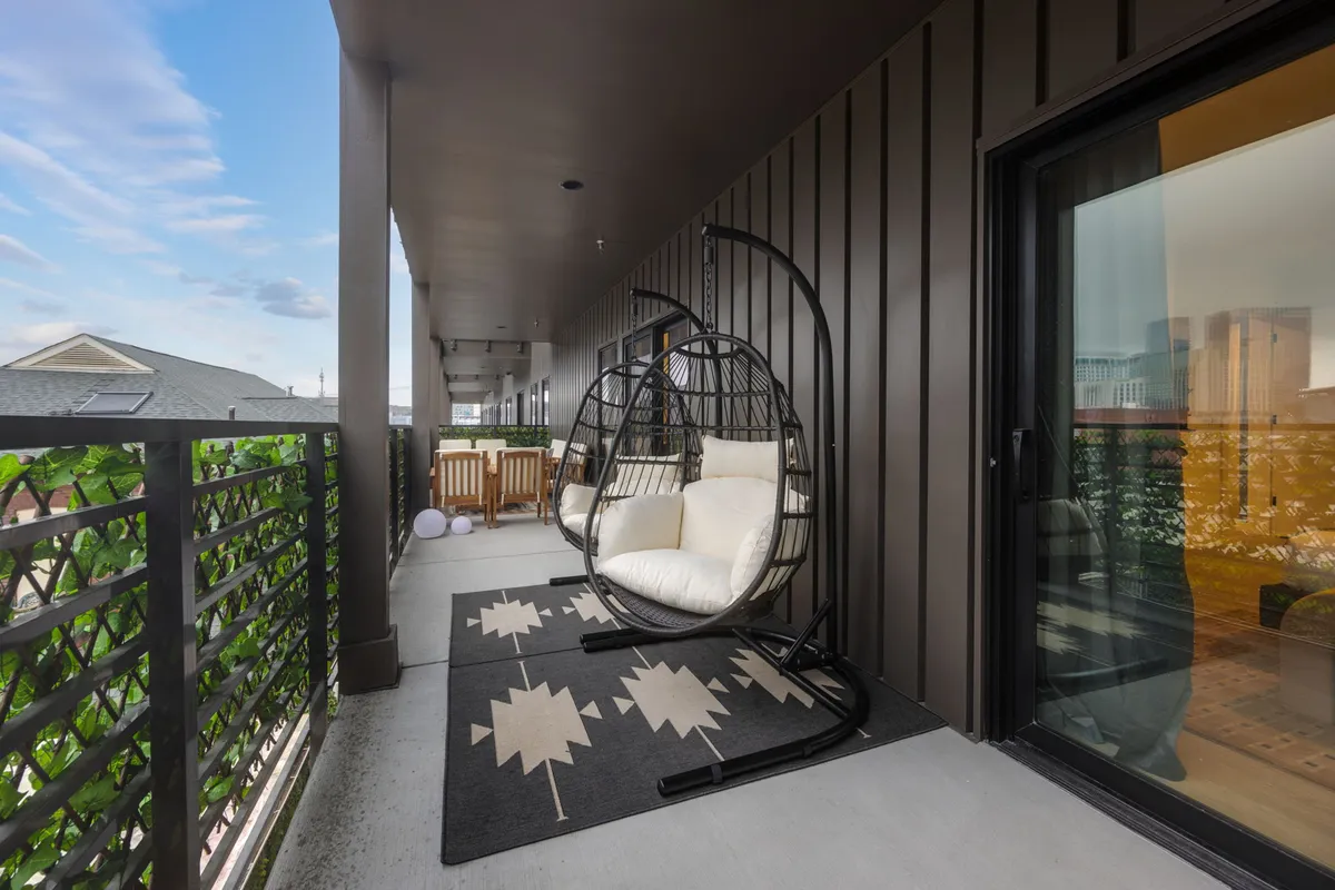 A calm and inviting balcony scene featuring a hanging chair, balcony railing, a cozy rug, and a captivating view of the city skyline in the background. The layout suggests a perfect spot for relaxing, reading, or enjoying the urban atmosphere with friends or a cup of coffee.