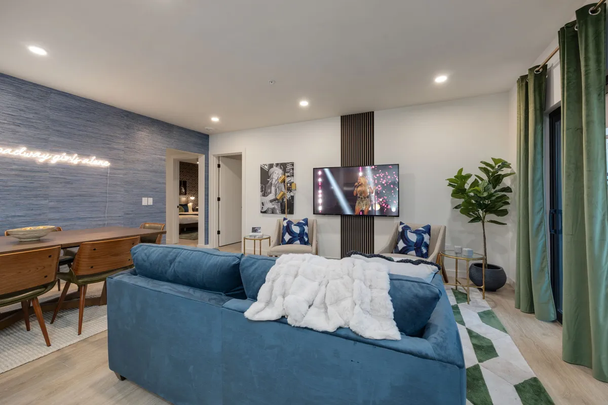 A cozy blue sofa faces a TV with a performance on screen, adjacent to a dining area with contemporary decor, green curtains, and a textured wall. The space exudes comfort and is ideal for movie nights, dinner parties, and social relaxation.