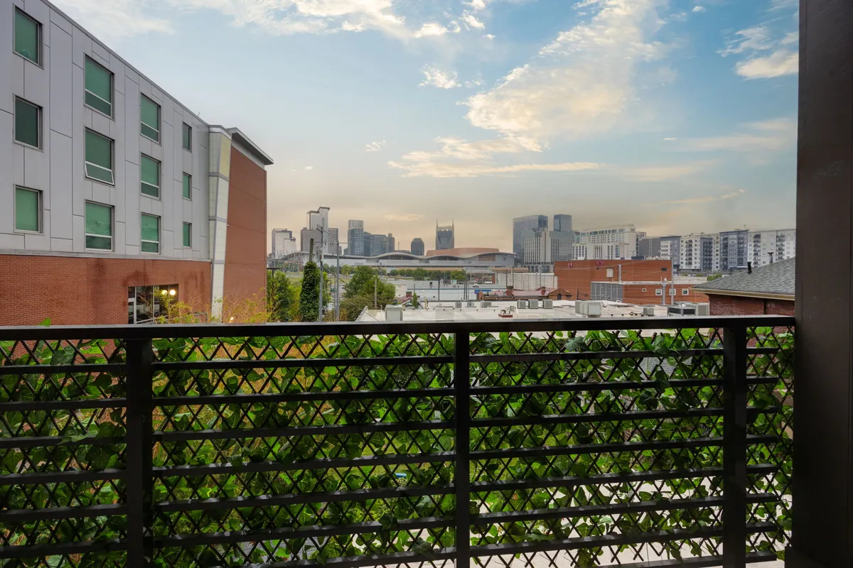 A perspective from the balcony overlooking the city skyline, with greenery draping along the balcony railing and clouds accenting the sky. The view is urban, open, and contemplative, perfectly capturing both daytime and sunset moods.