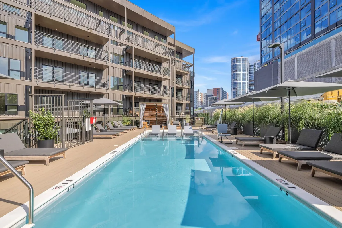 An inviting exterior shot featuring a sparkling swimming pool surrounded by lounge chairs, umbrellas, and well-tended landscaping. The building’s facade hints at urban living, while the pool area suggests a spot for pre-night-out relaxation or a morning recovery.
