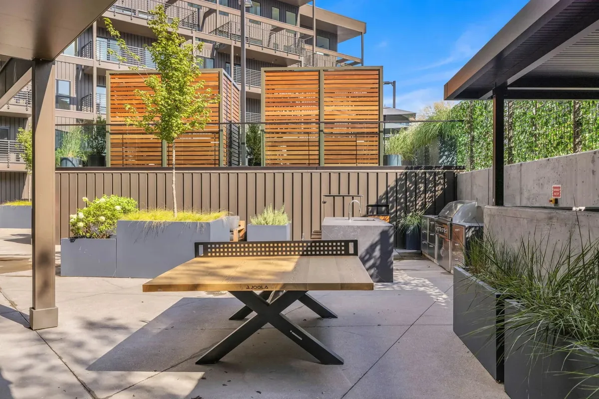 A welcoming outdoor BBQ and dining area, complete with a wooden table, lush greenery, decorative wooden panels, and a dedicated grilling station. The communal setup invites gatherings and memorable meals under the sky.