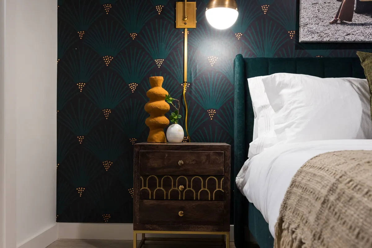 A stylish bedroom with a green velvet headboard, a decorative nightstand, and a stylish lamp. Soft natural light and tranquil decor create a restful, chic atmosphere.